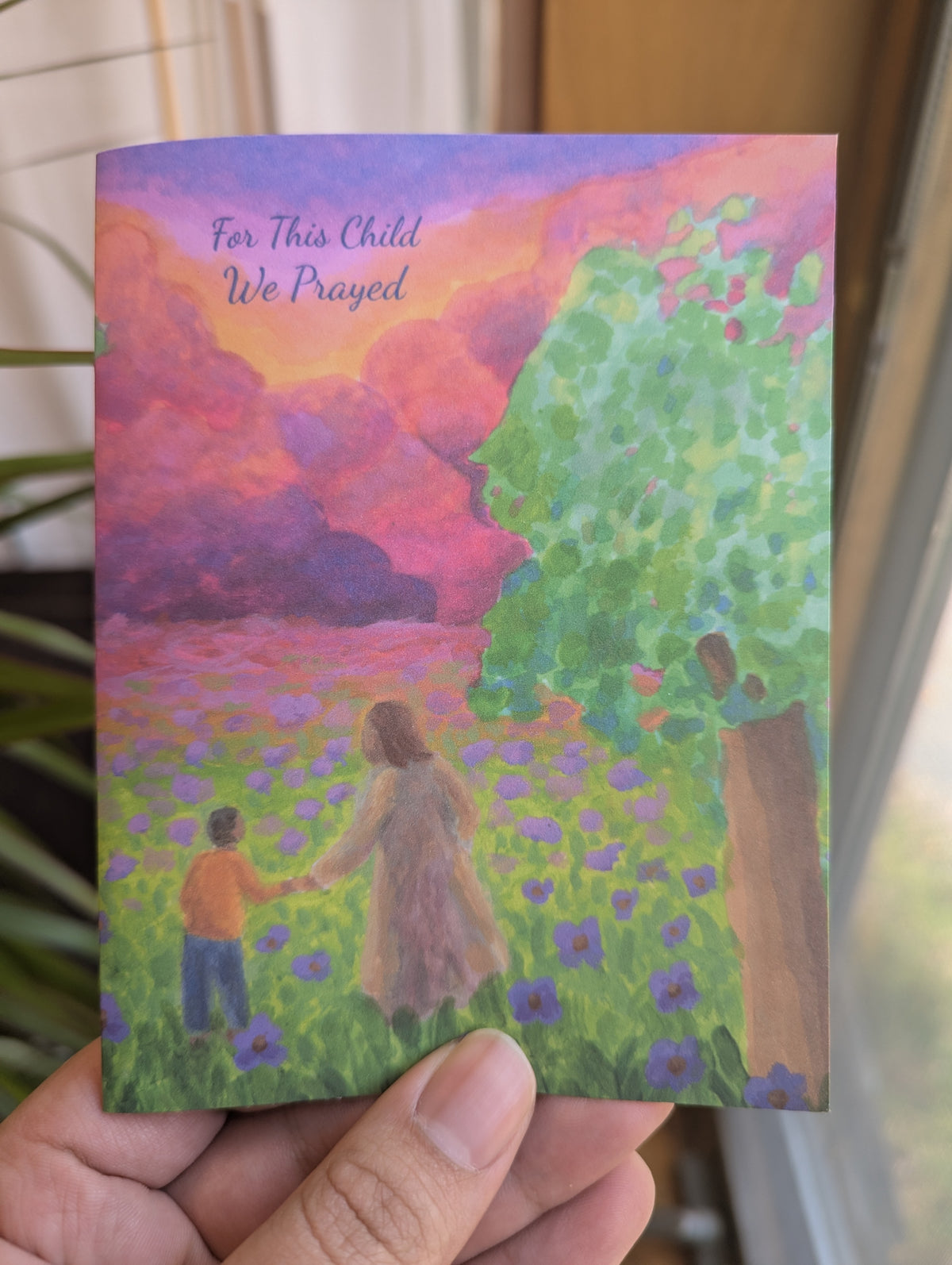 Christian baby dedication card with an original painting of Jesus holding a young child's hand, walking through a field of purple flowers toward a colorful sunset sky; cover reads "For This Child We Prayed"